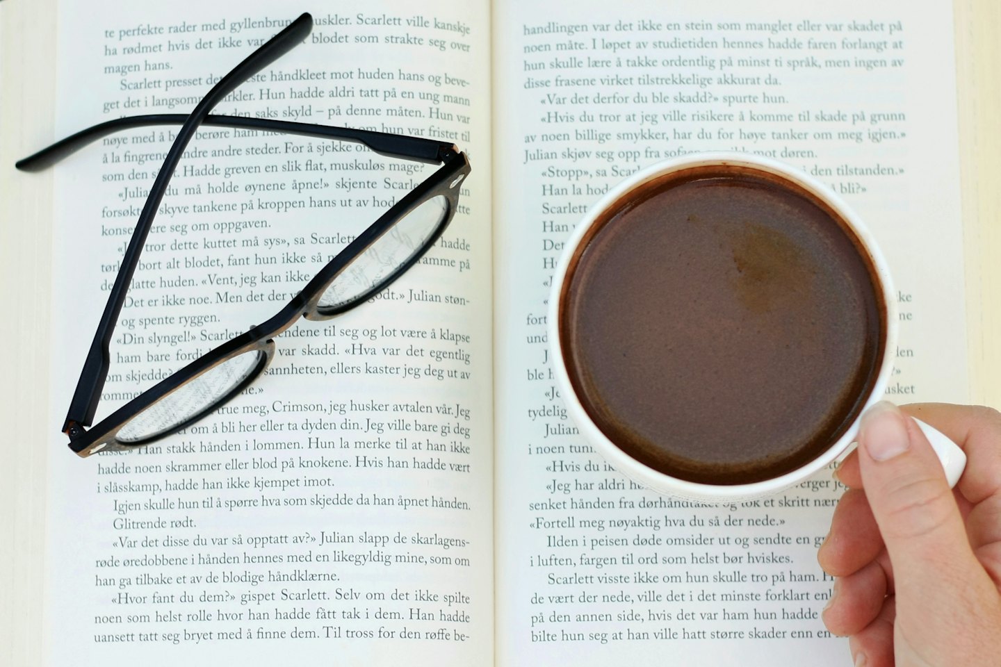 Open book with reading glasses and a hand holding coffee.